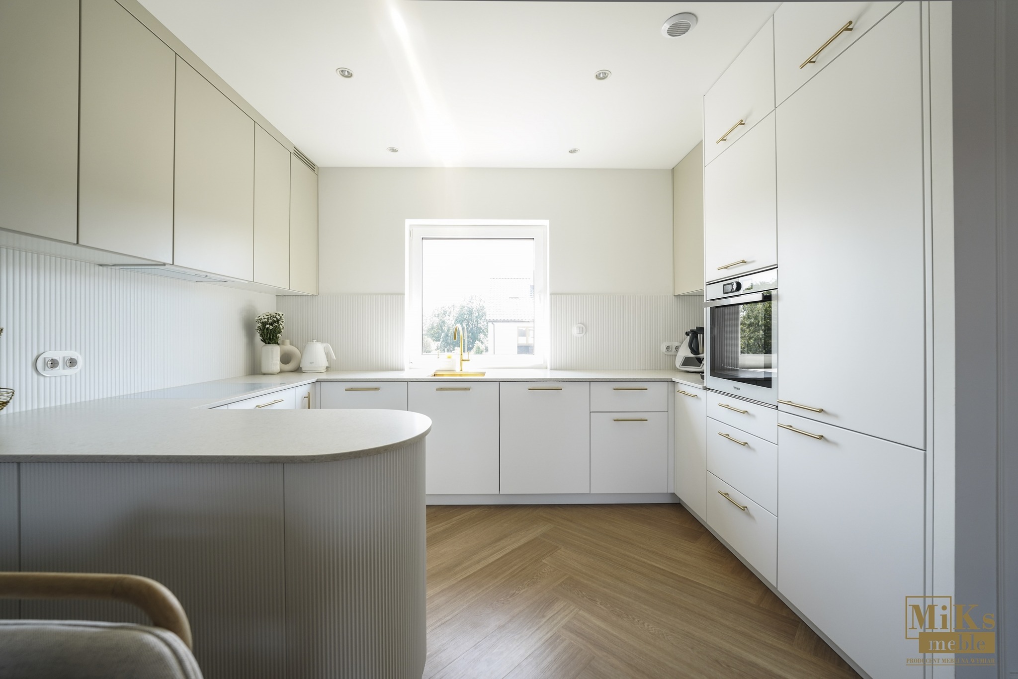 Jasna kuchnia z wyspą i złotymi uchwytami, parkiet w jodełkę, minimalistyczny design, okno z widokiem na zieleń, oświetlenie punktowe, frezowane fronty szafek.
