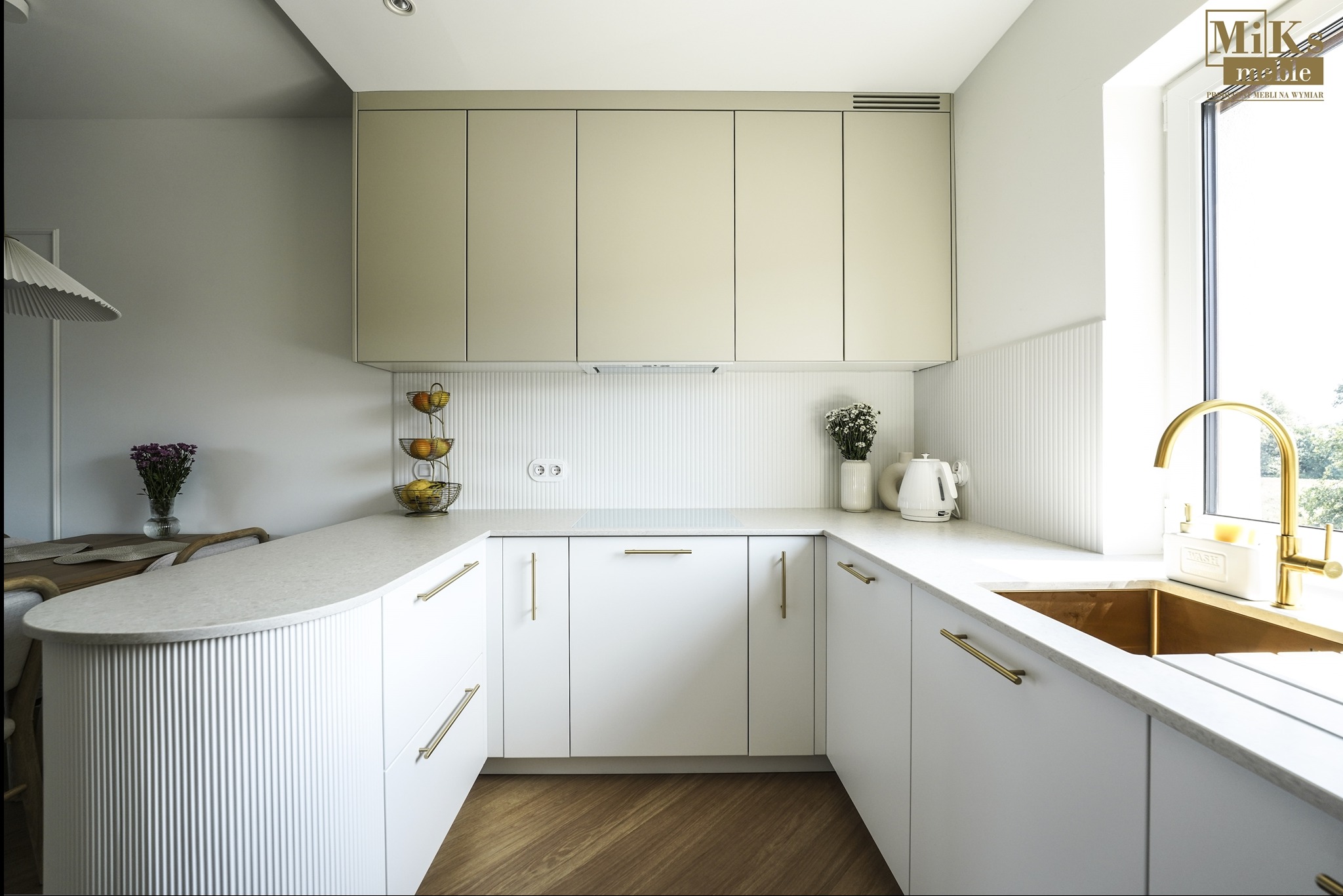 Jasna kuchnia w kształcie litery U z białymi frontami, złotymi uchwytami i blatem imitującym kamień. Uwagę przyciąga złota bateria i nietypowe wykończenie barku.