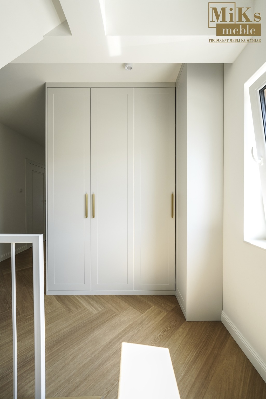Biała szafa wnękowa z trzema skrzydłami i złotymi uchwytami, w jasnym wnętrzu z parkietem ułożonym w jodełkę. Widok z góry, z fragmentem białej balustrady.