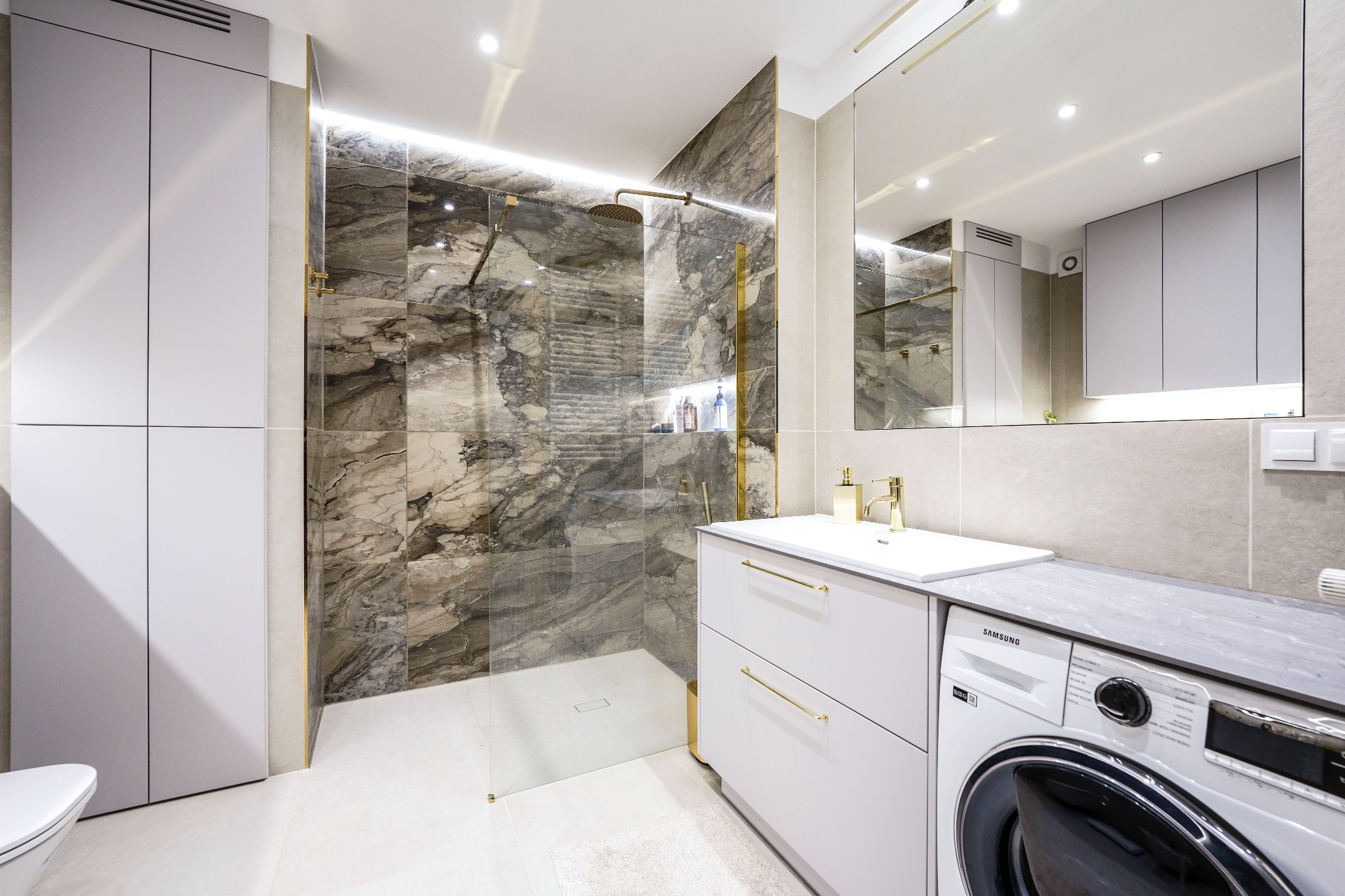 Nowoczesna łazienka z kabiną typu walk-in, marmurowymi płytkami i białymi szafkami z złotymi uchwytami, pralka Samsung widoczna w zabudowie.