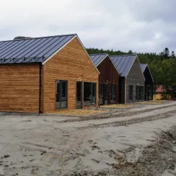 Rząd czterech nowoczesnych domów o prostej architekturze i drewnianej elewacji w różnych odcieniach brązu, z ciemnymi, skośnymi dachami i dużymi oknami, usytuowanych na rozległym, błotnistym...