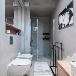 Minimalistyczna łazienka z betonowymi ścianami, wiszącą toaletą i bidetem, kabiną prysznicową ze szklanymi drzwiami oraz czarno-białym obrazem na ścianie.