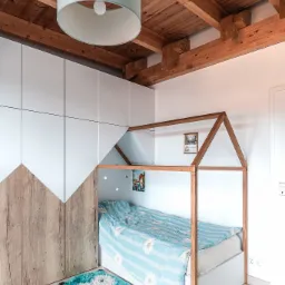 Pokój dziecięcy z łóżkiem w kształcie domku, szafą na wymiar z drewnianymi akcentami w kształcie gór i puszystym dywanikiem w kształcie sowy, oświetlony wiszącą lampą z drewnianym stropem z belkami.