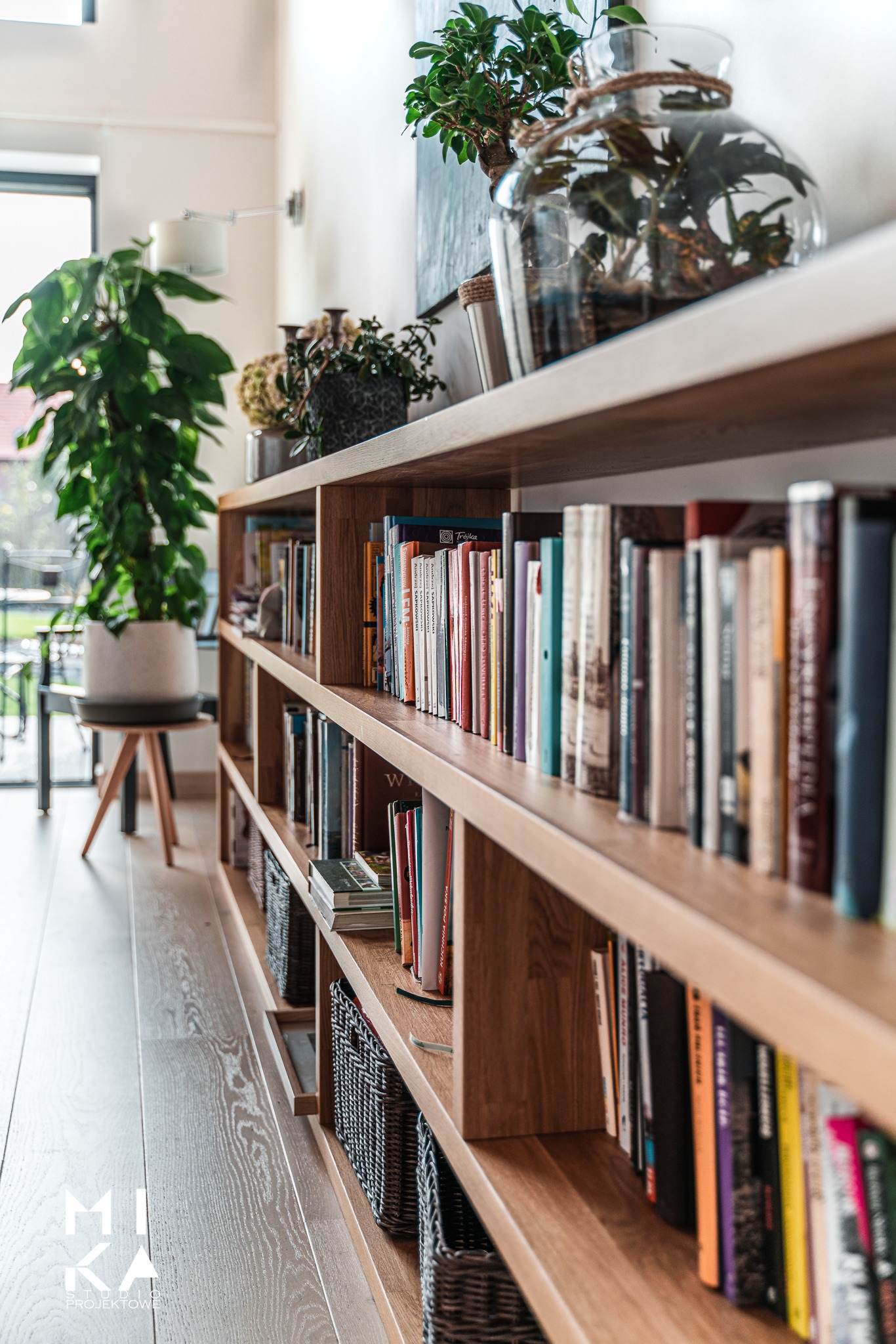 Regał z książkami i dekoracjami roślinnymi w jasnym wnętrzu, z widocznym fragmentem podłogi z jasnego drewna i logotypem projektowym w rogu.