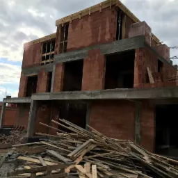 Wznoszenie dwukondygnacyjnego budynku z czerwonej cegły, widoczne betonowe elementy konstrukcyjne i drewniane szalunki, na pierwszym planie sterta odpadów drewnianych, w tle słup wysokiego napięcia.