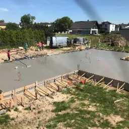 Wylewanie fundamentu pod budynek jednorodzinny, widoczna drewniana szalówka, świeżo wylany beton, zbrojenie wystające z betonu, ekipa budowlana w tle, dwa busy zaparkowane obok placu budowy.