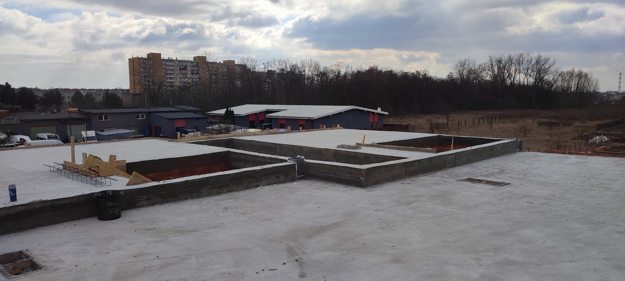 Wylany fundament z betonowymi ścianami szalunkowymi w trakcie budowy domu, widoczne zbrojenia i drewniane elementy szalunku, w tle zabudowa miejska i drzewa.