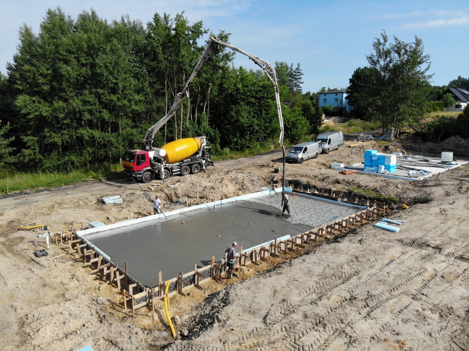 Wylewanie fundamentu pod budynek: beton z betonomieszarki podawany pompą, pracownicy rozprowadzają beton na zbrojeniu w szalunku, widoczne zaparkowane busy oraz materiały izolacyjne.