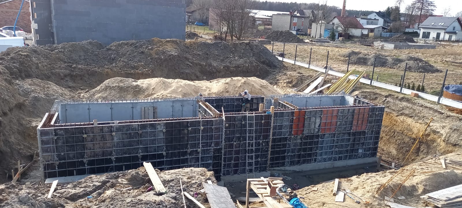 Wykop pod fundamenty z częściowo zaszalowanym i zazbrojonym betonem, widoczni pracownicy budowlani w trakcie pracy, w tle budynek w stanie surowym otwartym oraz zabudowa jednorodzinna.