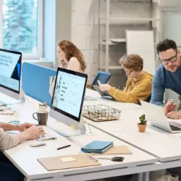 Zespół ludzi pracuje przy biurkach w jasnym biurze. Jeden mężczyzna w okularach pomaga kobiecie z dredami przy laptopie. Inni pracują na komputerach stacjonarnych i tabletach. Na biurkach widoczne...