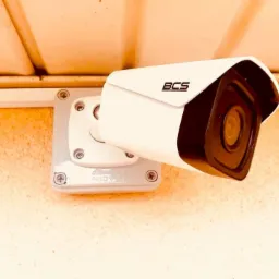 Biała kamera monitoringu BCS zamontowana na szarej podstawie z widocznymi śrubami, przymocowana do beżowej ściany pod jasnym sufitem.