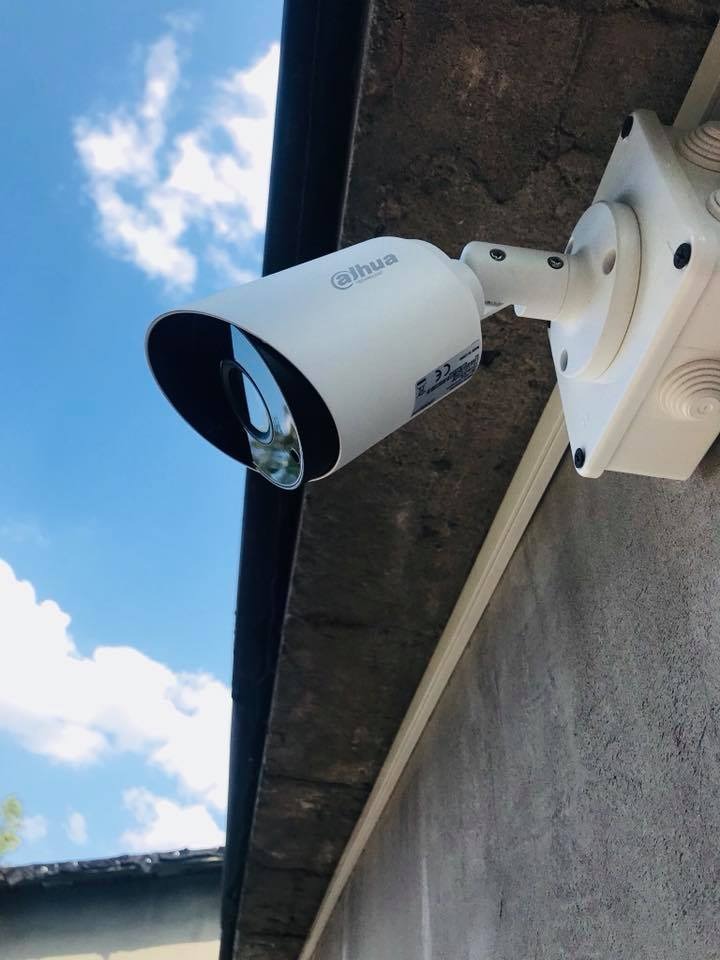 Biała kamera monitoringu Dahua zamontowana na szarej ścianie budynku pod błękitnym niebem z chmurami, widoczna puszka instalacyjna.