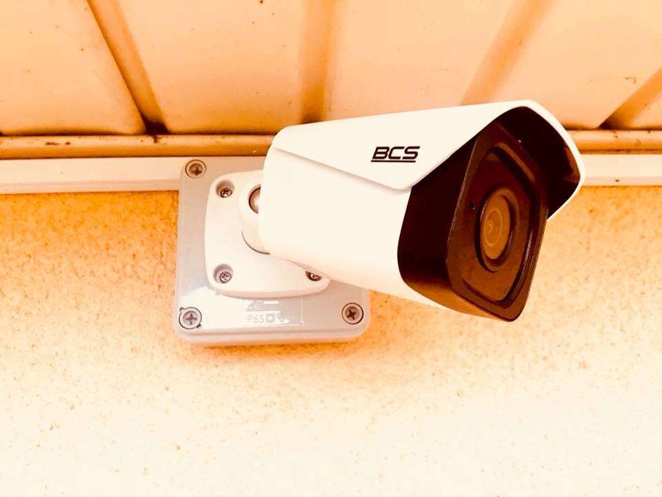 Biała kamera monitoringu BCS zamontowana na szarej podstawie z widocznymi śrubami, przymocowana do beżowej ściany pod jasnym sufitem.