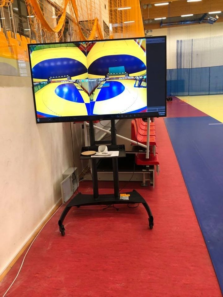 Monitor na mobilnym stojaku wyświetla obraz z kamer monitoringu hali sportowej z widocznym boiskiem do piłki ręcznej.