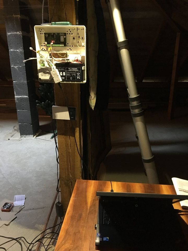 Otwarta skrzynka centrali alarmowej z akumulatorem na drewnianym słupie w surowym pomieszczeniu, widoczne narzędzia i laptop na biurku, instalacja w trakcie podłączania.