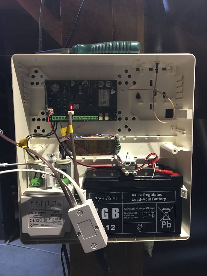 Otwarta obudowa centrali alarmowej z widoczną płytą główną, akumulatorem awaryjnym i transformatorem, zielony śrubokręt leżący na górnej krawędzi obudowy.
