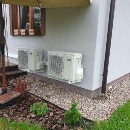 PHU MARTIV Mariusz Żuczkowski - Dwa klimatyzatory AUX zamontowane na ścianie domu z szarą elewacją, widoczne kamienie ozdobne i trawa, zadaszenie parasola w tle.