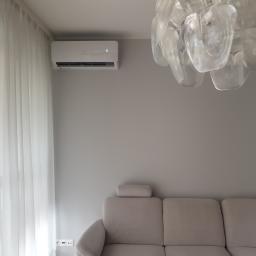 PHU MARTIV Mariusz Żuczkowski - Nowoczesny klimatyzator ścienny z wyświetlaczem temperatury, zamontowany w jasnym salonie z beżową sofą i designerskim żyrandolem z tworzywa.