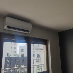 PHU MARTIV Mariusz Żuczkowski - Biały klimatyzator marki AUX zamontowany na ścianie w mieszkaniu z widokiem na nowoczesne budynki przez okno.