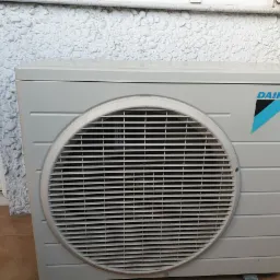 Zewnętrzna jednostka klimatyzacji Daikin zamontowana przy ścianie budynku o nierównej, białej fakturze. Widoczny fragment okna po lewej stronie.