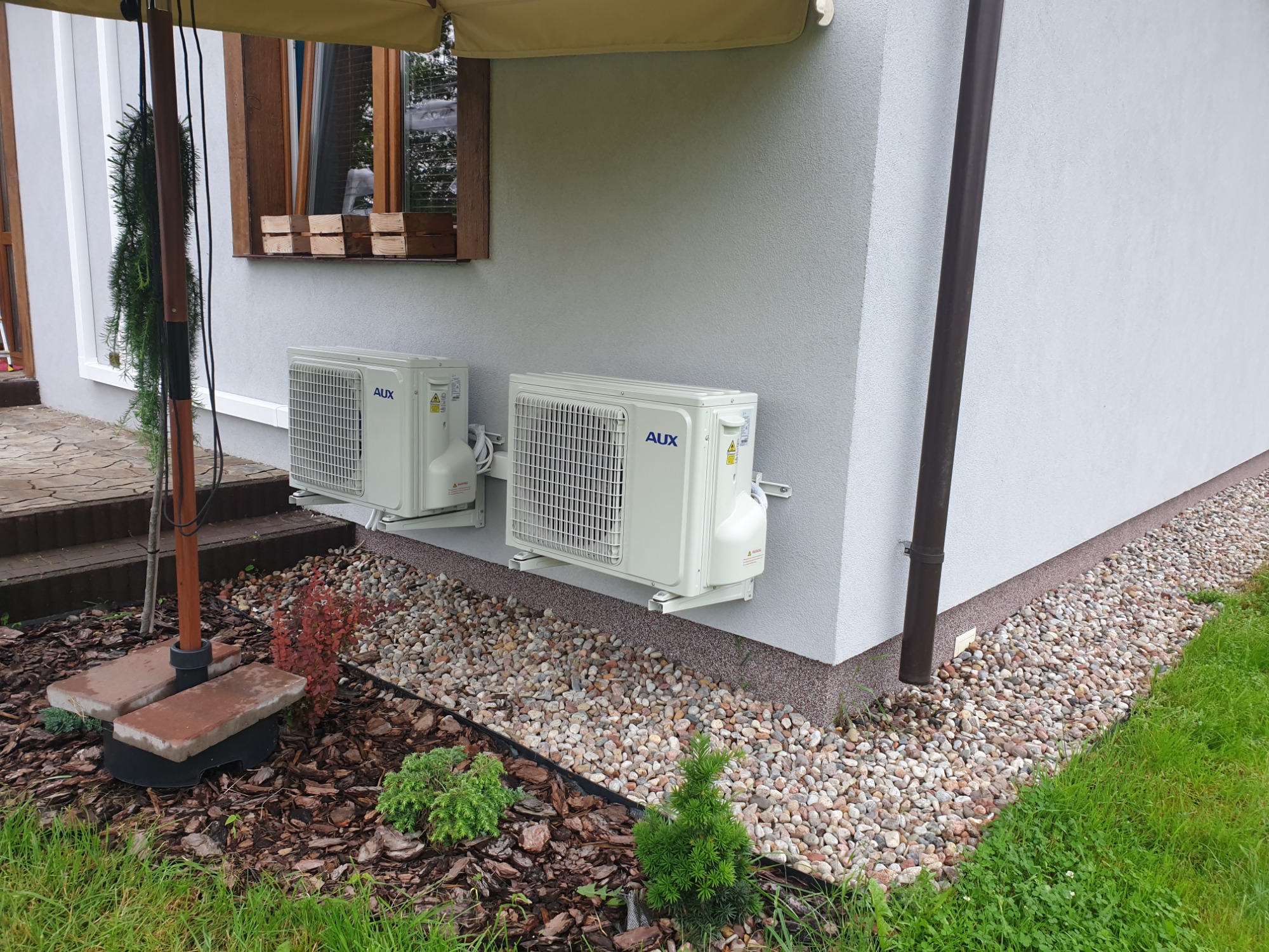 Dwa klimatyzatory AUX zamontowane na ścianie domu z szarą elewacją, widoczne kamienie ozdobne i trawa, zadaszenie parasola w tle.