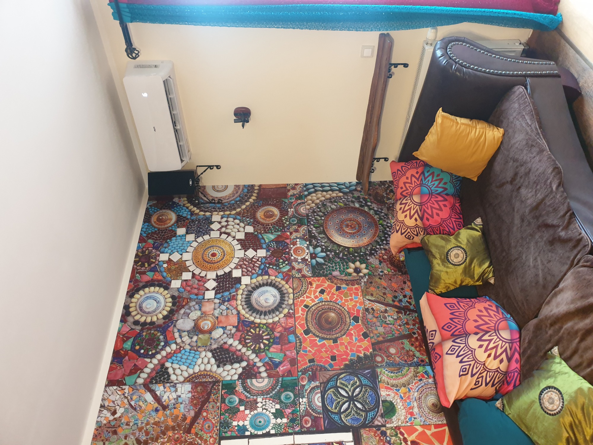 Widok z góry na wnętrze pokoju z zamontowanym klimatyzatorem ściennym AUX, wzorzystą mozaikową podłogą, brązową sofą i dekoracyjnymi poduszkami w odcieniach żółci, zieleni i pomarańczy.