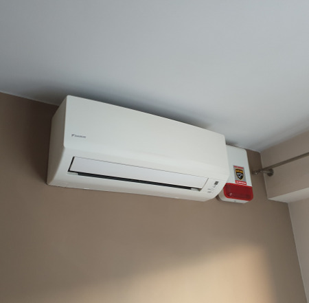 Biały klimatyzator Daikin zamontowany na ścianie w pomieszczeniu, obok czujnik dymu z czerwonym światłem alarmowym.