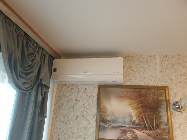 Biały klimatyzator LG zamontowany na ścianie z tapetą o wzorze marmurkowym, obok wiszą zasłony i obraz w złotej ramie.