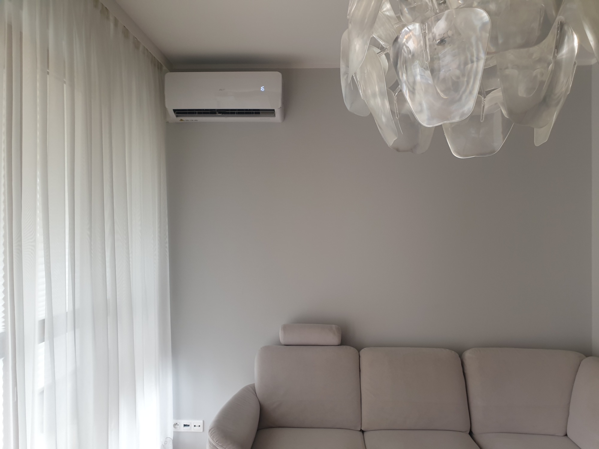 Nowoczesny klimatyzator ścienny z wyświetlaczem temperatury, zamontowany w jasnym salonie z beżową sofą i designerskim żyrandolem z tworzywa.
