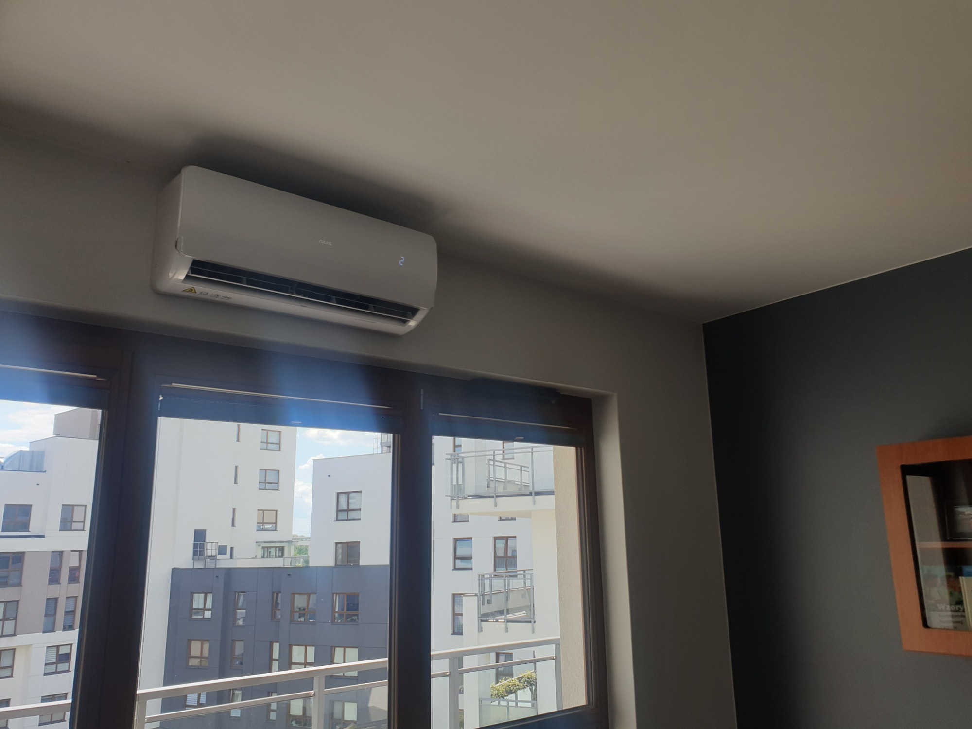 Biały klimatyzator marki AUX zamontowany na ścianie w mieszkaniu z widokiem na nowoczesne budynki przez okno.