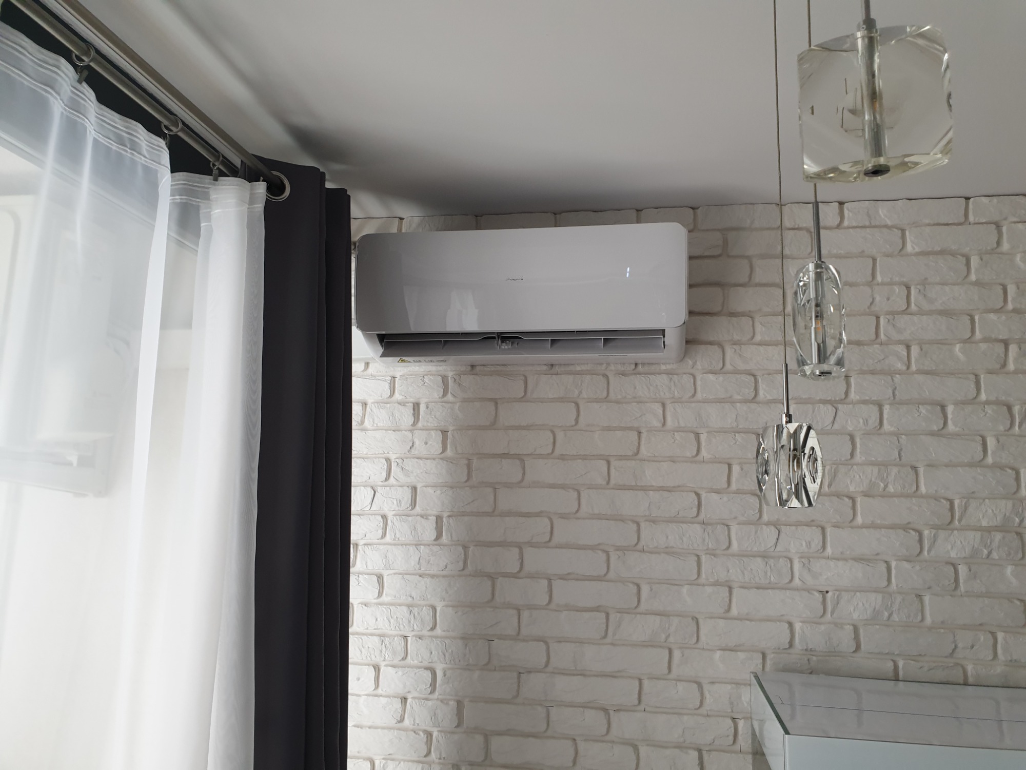 Biały klimatyzator na białej ceglanej ścianie, obok okna z zasłonami i ozdobnych lamp wiszących.
