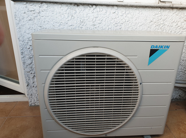 Zewnętrzna jednostka klimatyzacji Daikin zamontowana przy ścianie budynku o nierównej, białej fakturze. Widoczny fragment okna po lewej stronie.