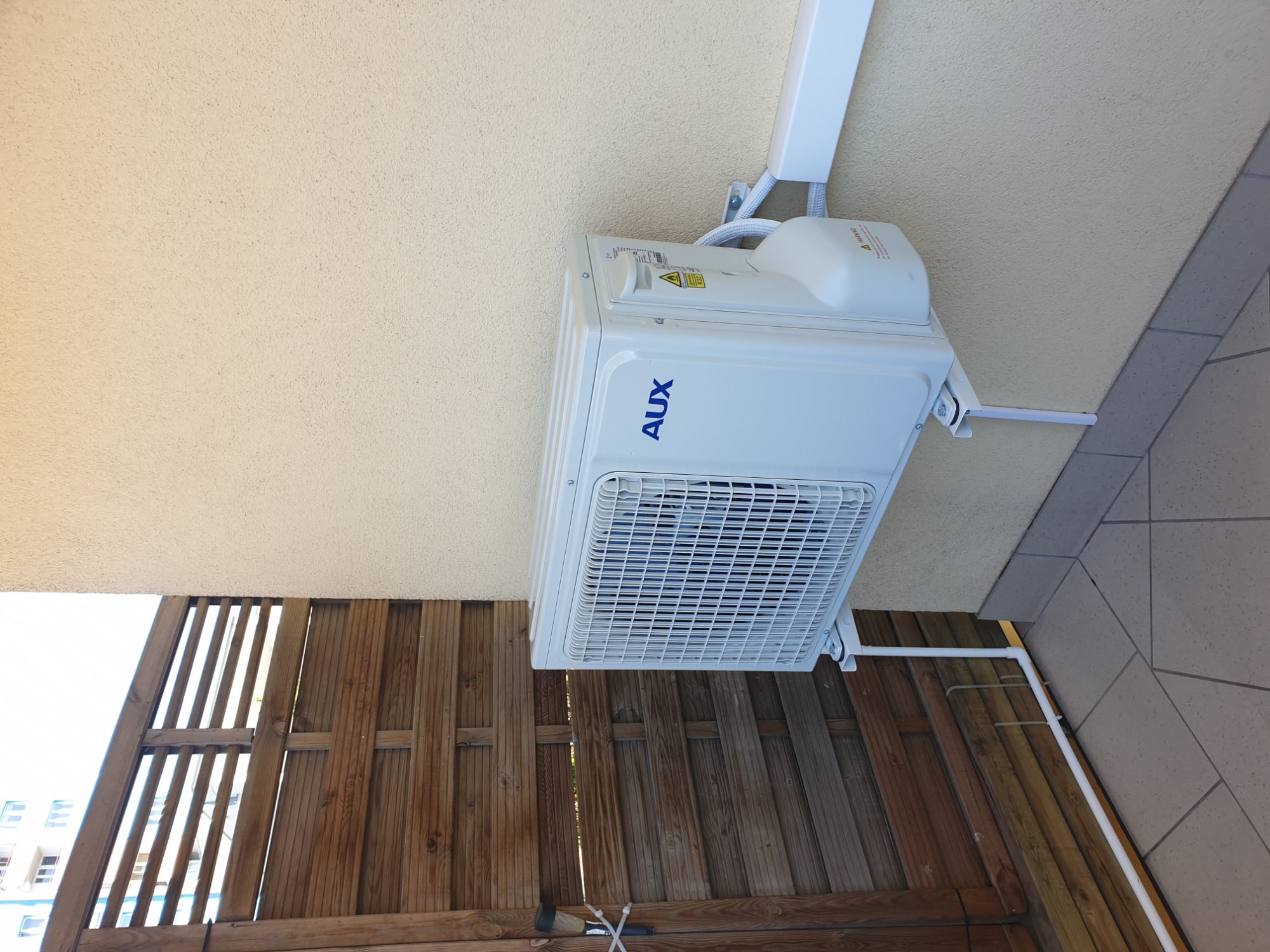 Zewnętrzna jednostka klimatyzatora marki AUX zamontowana na elewacji budynku, widoczna z perspektywy balkonu z drewnianą balustradą.