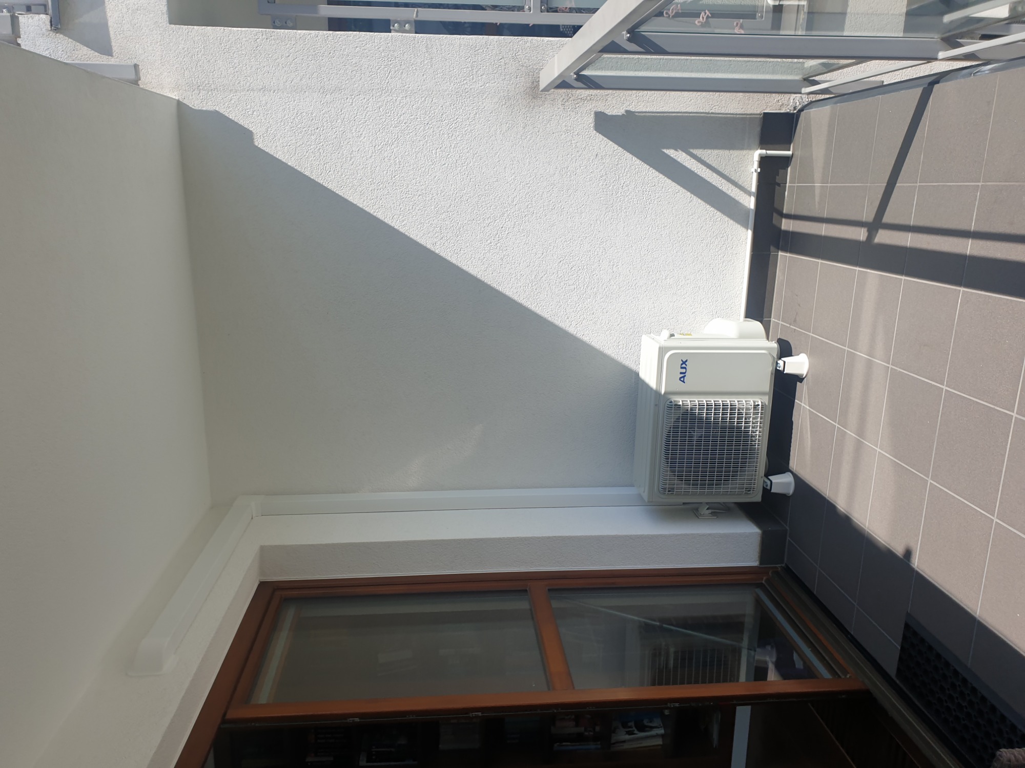 Biała jednostka zewnętrzna klimatyzatora AUX zamontowana na balkonie, widoczna z góry pod kątem, z fragmentem okna i płytek balkonowych w tle.