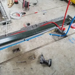 Betonowa podłoga w trakcie prac instalacyjnych, widoczne rury w kolorach czerwonym, niebieskim i żółtym, wiertarka akumulatorowa leży obok wywierconego otworu.