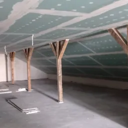 Poddasze w trakcie montażu płyt karton-gips na skośnym suficie, widoczne drewniane słupy podtrzymujące konstrukcję dachu, fragmenty płyt na podłodze i worki z odpadami.