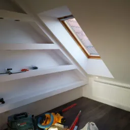 Poddasze w Pelplinie po remoncie: wnęki w ścianie z oświetleniem, okno dachowe z widokiem na dachówkę, grzejnik, na podłodze narzędzia i odkurzacz.