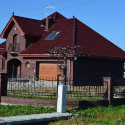 Dwukondygnacyjny dom z ciemnej cegły z czerwoną dachówką, ozdobnym balkonem i kutym ogrodzeniem. Widoczny garaż z drewnianą bramą i dekoracyjne oświetlenie zewnętrzne.