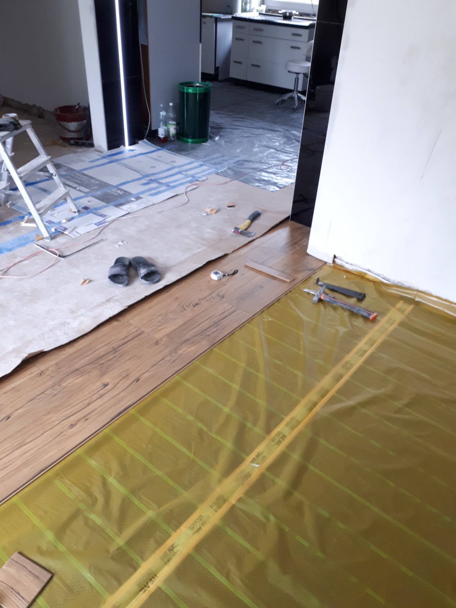 Układanie paneli podłogowych na folii grzewczej, widoczne narzędzia, młotek i przymiar, w tle drabina i resztki materiałów budowlanych, na podłodze leżą klapki.