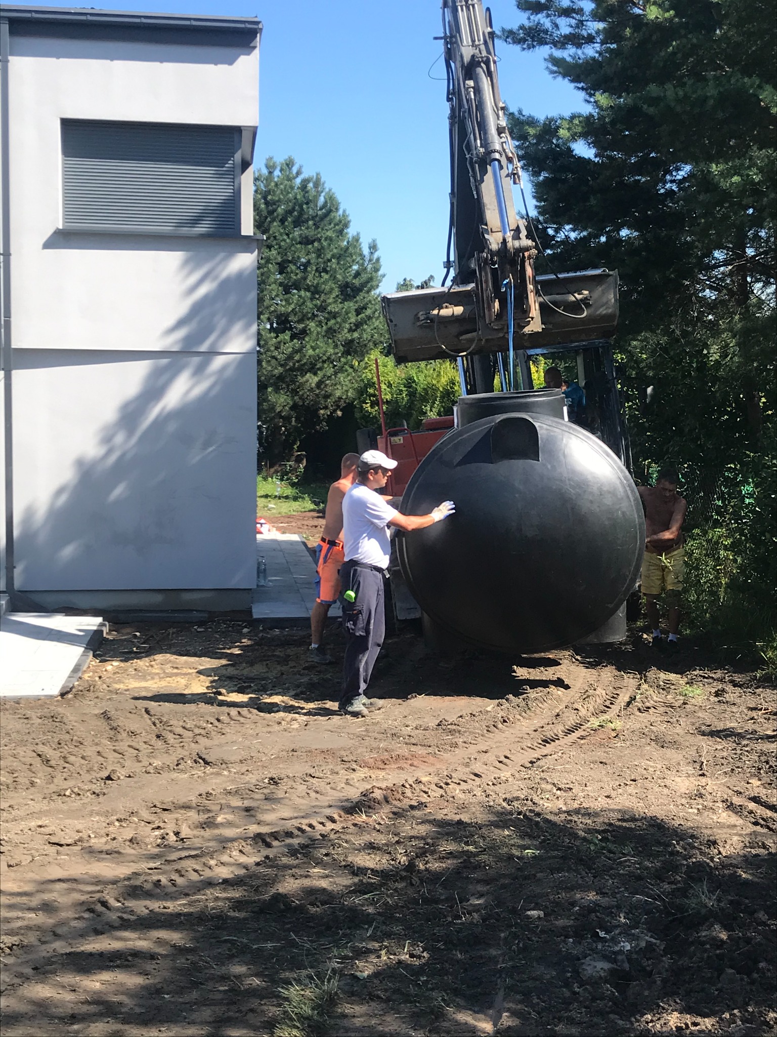 Montaż szamba: Czarny, kulisty zbiornik umieszczany za pomocą koparki na przygotowanym terenie obok nowoczesnego domu z szarą elewacją i roletami zewnętrznymi, trzech pracowników nadzoruje proces.