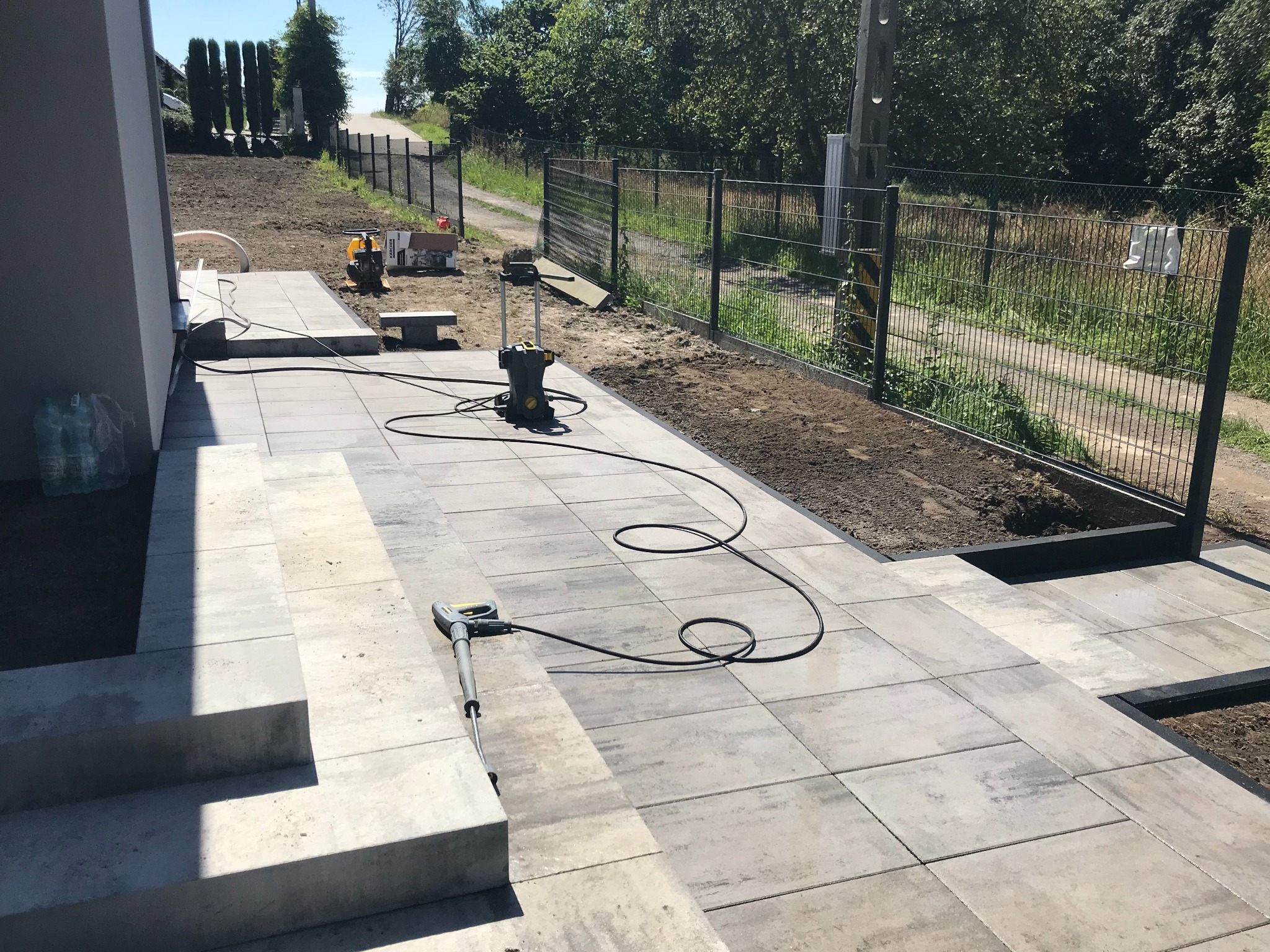 Nowo ułożony taras z kostki brukowej w odcieniach szarości prowadzący do furtki ogrodowej, z widoczną myjką ciśnieniową Karcher i ubijarką wibracyjną na dalszym planie.