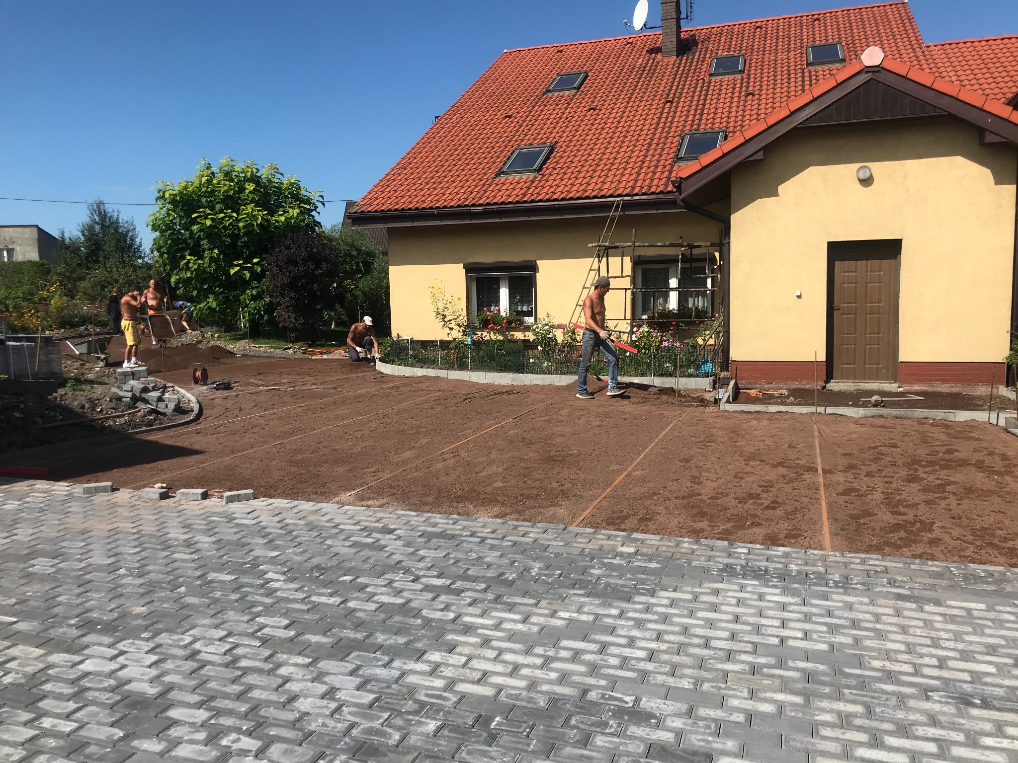 Ekipa budowlana przygotowuje teren pod posesją z żółtym domem i czerwoną dachówką, widoczne częściowo ułożone szare kostki brukowe oraz wyrównana ziemia, mężczyzna w pomarańczowej kamizelce...