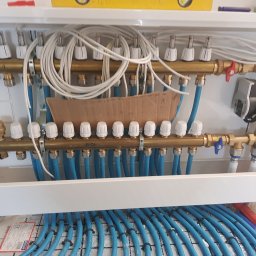 Instalacje Maciej Pędowski - Rozdzielacz ogrzewania podłogowego w szafce instalacyjnej, widoczne liczne pętle rur w kolorze niebieskim, białe termostaty na górnej belce rozdzielacza, pompa obiegowa i zawory odcinające...