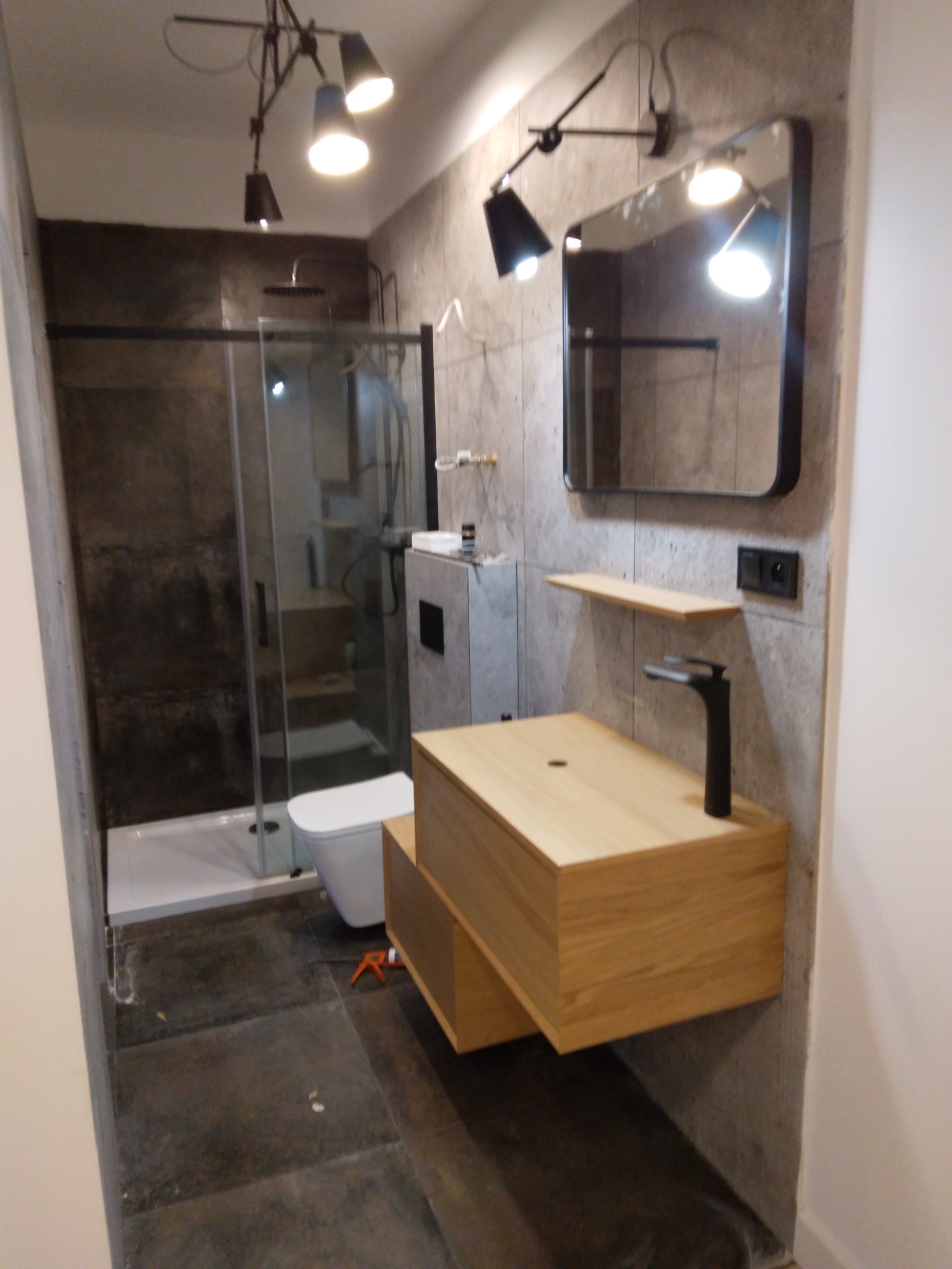 Nowoczesna łazienka z kabiną prysznicową typu walk-in, wiszącą szafką z umywalką z jasnego drewna i czarną armaturą, oświetlona lampami sufitowymi w stylu industrialnym, z widocznym silikonem...