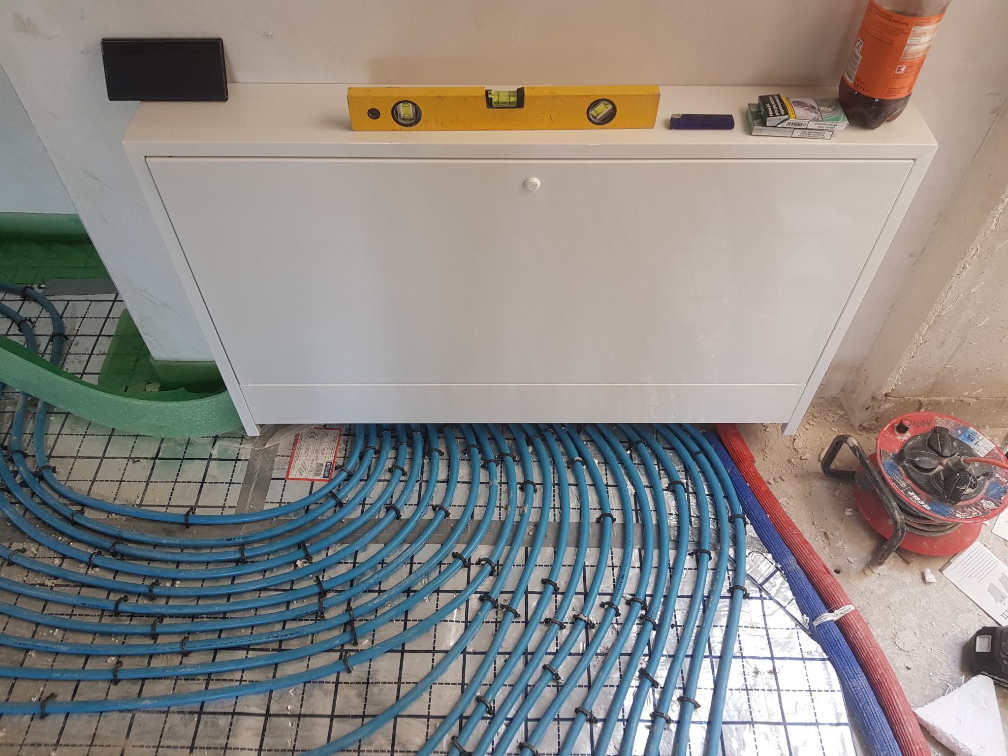 Rozdzielacz ogrzewania podłogowego w białej szafce, z widocznymi niebieskimi rurami pex mocowanymi do maty systemowej, poziomica na szafce, widoczny fragment przewodu elektrycznego na bębnie.