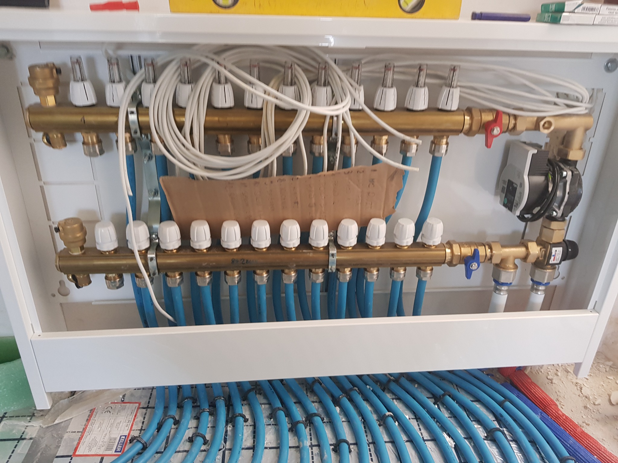 Rozdzielacz ogrzewania podłogowego w szafce instalacyjnej, widoczne liczne pętle rur w kolorze niebieskim, białe termostaty na górnej belce rozdzielacza, pompa obiegowa i zawory odcinające...