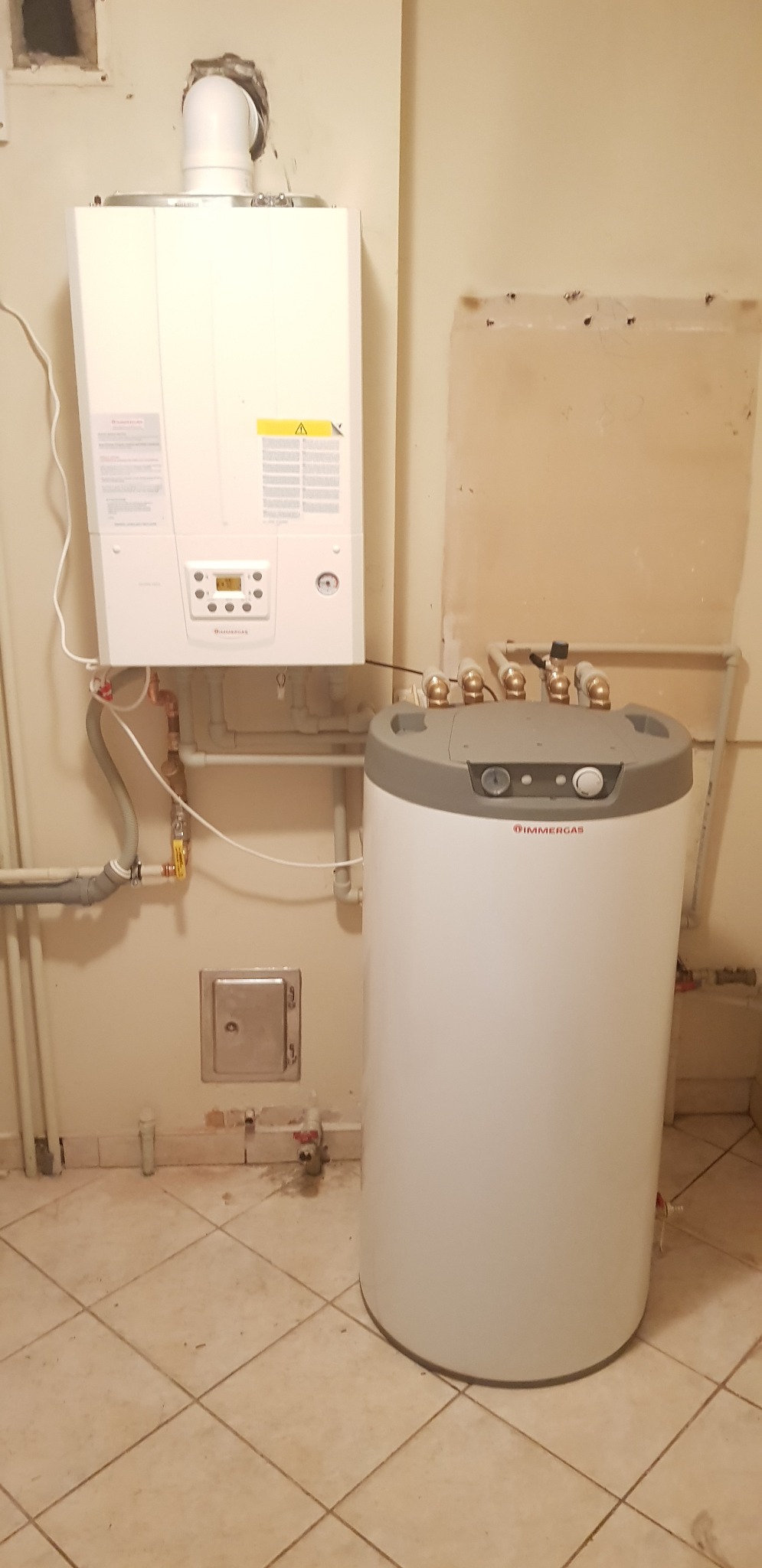 Biały piec gazowy Immergas wiszący na ścianie, obok biały zasobnik ciepłej wody z szarym wykończeniem, widoczne rury i przyłącza, tło stanowi beżowa ściana i podłoga z płytek.
