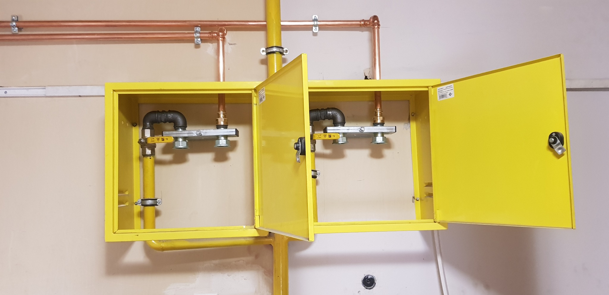 Żółta szafka gazowa z otwartymi drzwiczkami, widoczne instalacje miedziane i gazowe z zaworami, zamontowana na beżowej ścianie.