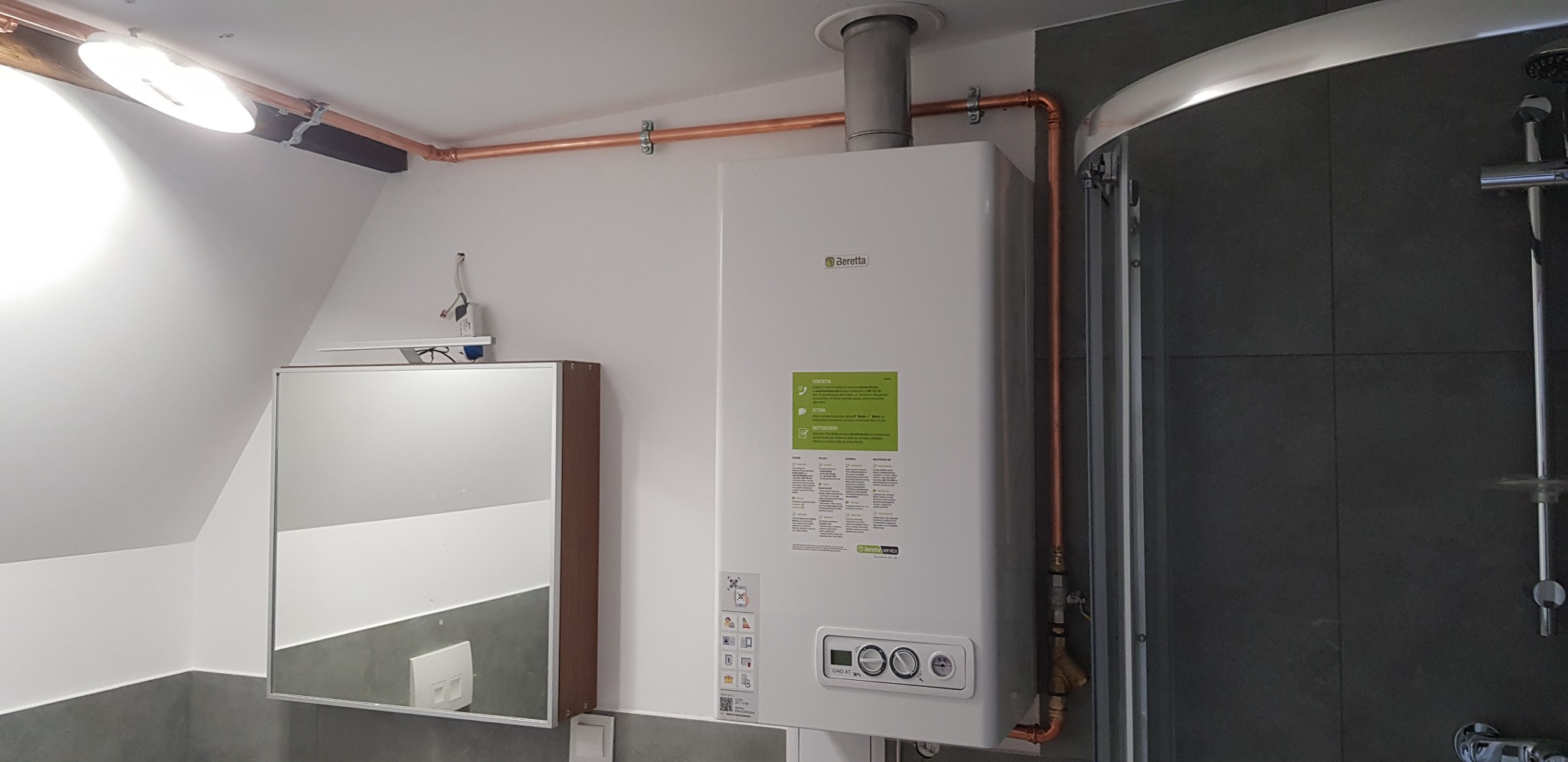 Biały piec gazowy Beretta zamontowany w łazience z miedzianymi rurami, widoczna kabina prysznicowa z szarymi płytkami i lustro z szafką.