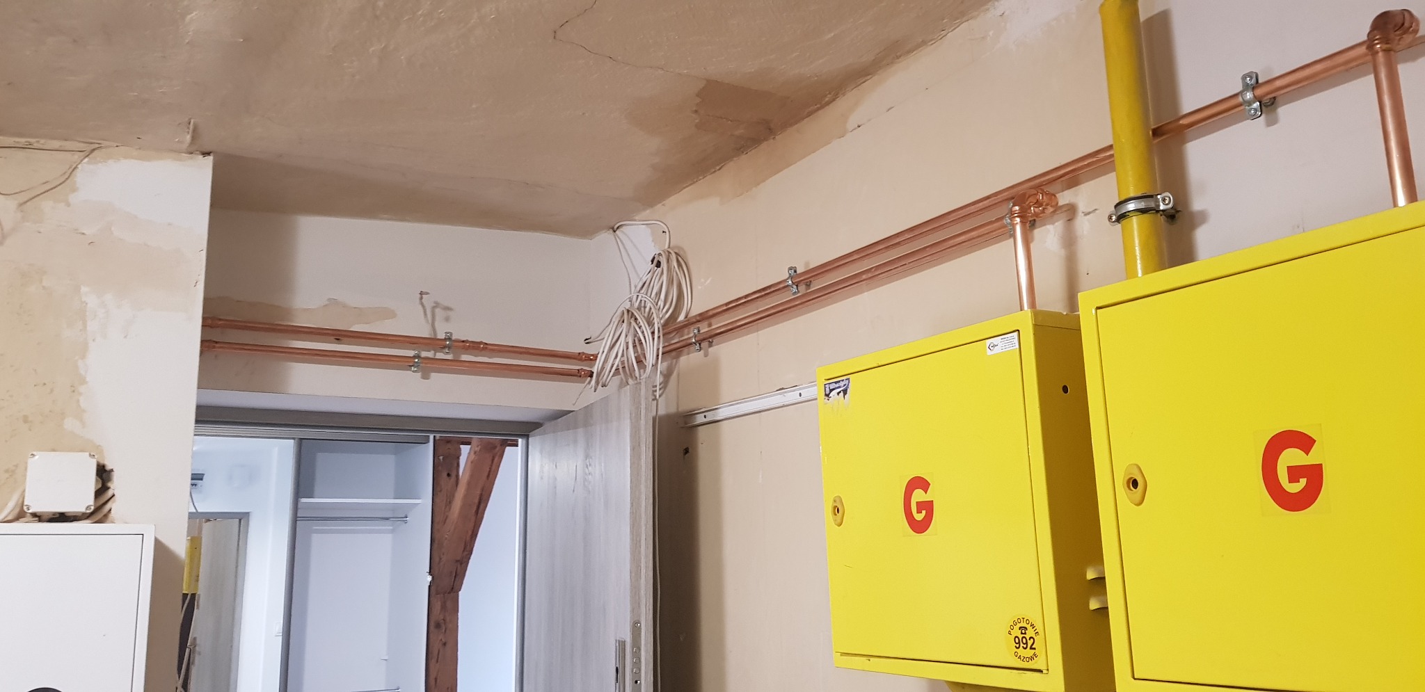 Żółte skrzynki gazowe z czerwonym logo G, miedziane rury instalacji gazowej zamontowane w pomieszczeniu w Poznaniu, widoczne kable i niedokończone prace wykończeniowe.